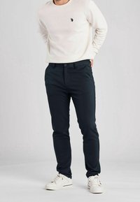 Námořnické kalhoty se slim fit, vyrobené z elastického materiálu; mají zapínání na knoflík a boční kapsy, v kombinaci s světlým krémovým svrškem.