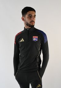 Chaqueta deportiva negra con detalles en dorado, cuenta con rayas rojas y azules en los hombros, cuello con cremallera y el logo del Olympique de Lyon en el pecho.