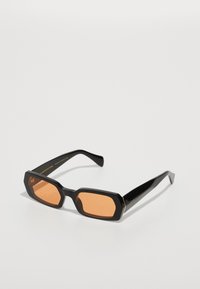 Gafas de sol negras con un diseño angular y lentes naranjas. La montura es brillante con una textura suave, con un sutil branding en la patilla.