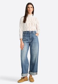 Donna posa con sicurezza in una blusa di pizzo bianca e jeans a vita alta, con le mani nelle tasche. Indossa scarpe beige casual. Sfondo semplice.