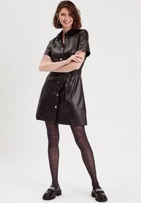 Robe chemise en cuir noir avec des manches courtes, des boutons sur le devant et une ceinture à nouer. Portée avec des collants à motifs sombres et des mocassins épais.