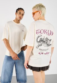 Krém oversized pólók lila "TOKYO SHINJUKU" grafikával és tigrist ábrázoló mintával a hátsó oldalon. Farmer rövidnadrággal és fekete szoknyával viselve.
