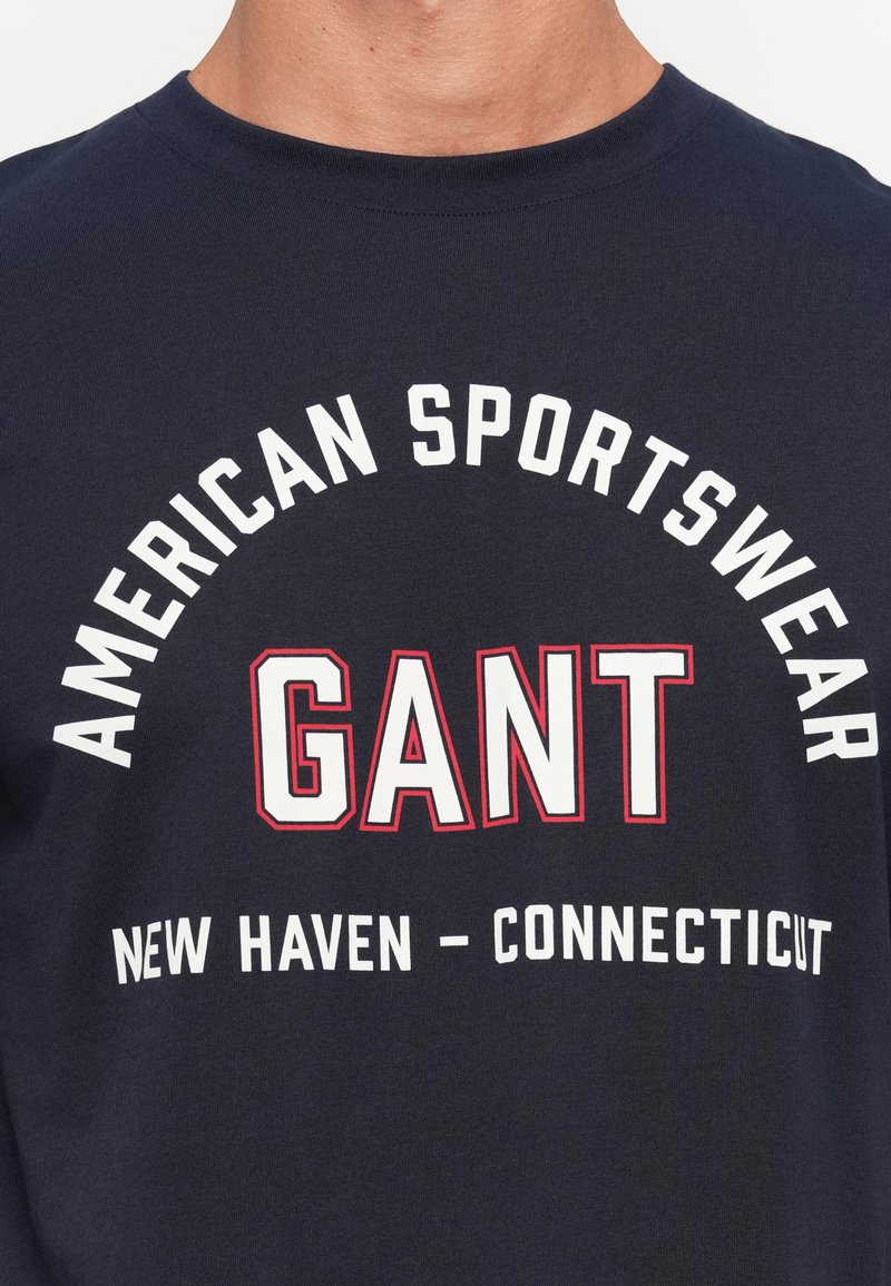 Granatowa bawełniana koszulka z pogrubionym białym i czerwonym napisem: "AMERICAN SPORTSWEAR GANT NEW HAVEN - CONNECTICUT" w zakrzywionym układzie.
