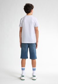 Vit bomullströja, blå denimshorts, vita sneakers med gröna detaljer och vita strumpor med blå ränder, sedd bakifrån.