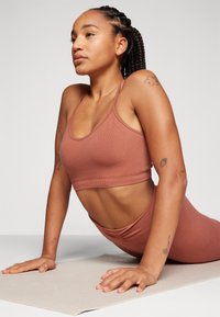 Brun ribbet sportstopp med tynne stropper, kombinert med matchende høytaljede bukser, brukt mens man er i en yoga posisjon på en matte.