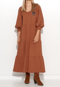 Robe brune à volants en tissu doux, dotée d'un col rond et de manches bouffantes. Détail de badge brodé sur la poitrine. Portée avec des bottines brunes.