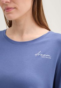 Sininen puuvillapaita, jossa on pyöreä kaula-aukko ja vasemmalla puolella valkoinen "dream. TOM TAILOR" -logo. Sileä rakenne, rento istuvuus.