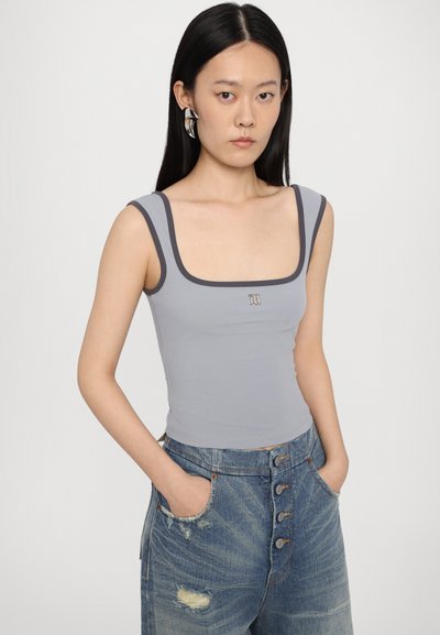 Grå tætsiddende tanktop med bred firkantet udskæring, kontrasterende mørk kant og lille logoaccent på brystet; parret med slidte blå jeans.