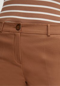 Pantalon marron sur mesure avec une texture lisse, doté d'une fermeture à bouton et de poches latérales. Associé à un haut rayé marron et blanc.