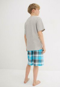 T-shirt en coton gris associé à un short à carreaux bleus avec des accents noir et blanc. Modèle vu de dos, pieds nus.
