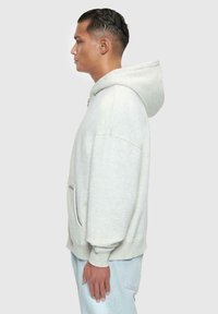 Lys grå hoodie med en frontlomme, snøre til hætten og ribbede ærmer, med et afslappet fit og blød tekstur. Model vist i profil.