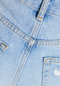 Vaaleansininen farkkukangas, jossa on tekstuuri, oranssit ompeleet, näkyvät saumat ja tasku repaleisilla reunoilla.