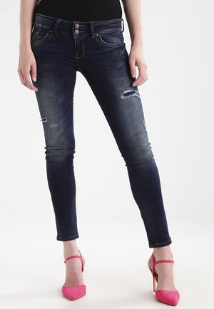 Kvinde iført mørkeblå, slidte, tætsiddende jeans og lyserøde højhælede sko, stående mod en hvid baggrund.