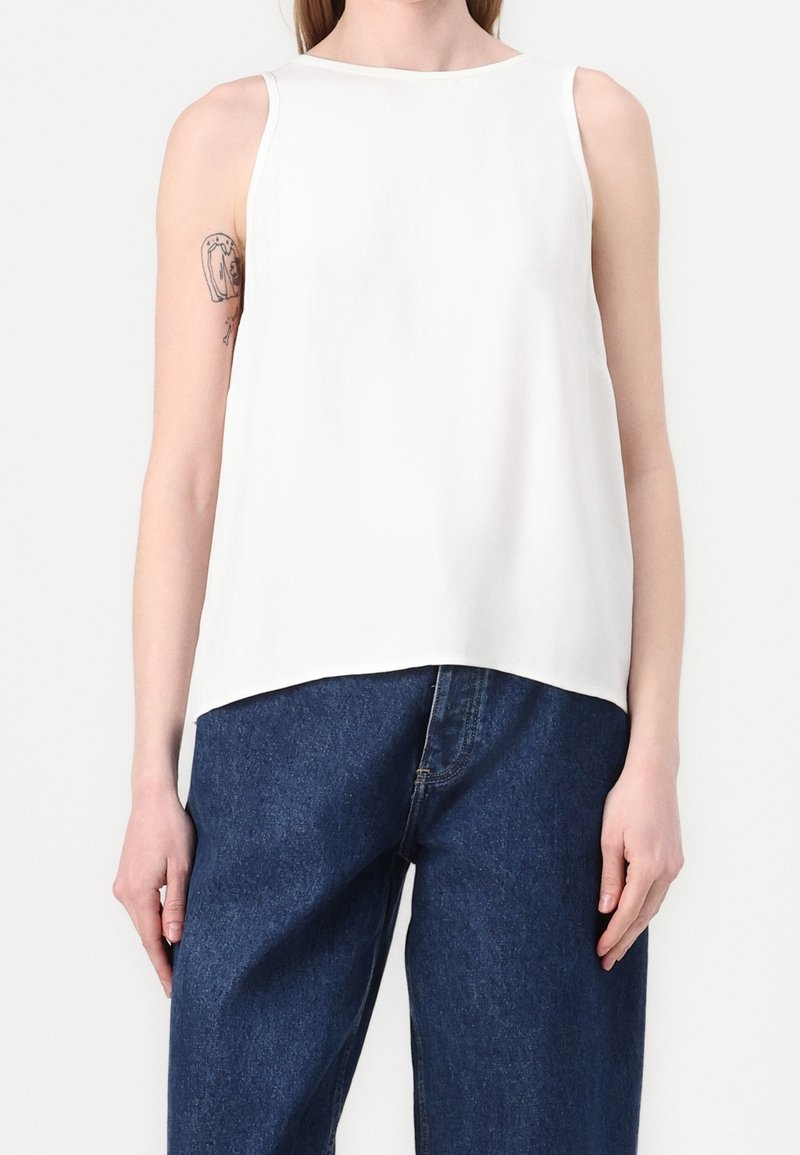 Femme portant un haut blanc sans manches et un jean large bleu foncé, avec un tatouage visible sur le haut du bras gauche, debout sur un fond blanc.