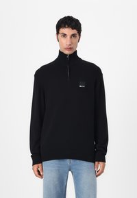 Maglione nero a maglia con colletto alto e chiusura a mezzo zip sul davanti. Presenta un'etichetta quadrata sul petto e polsini e orlo a coste.