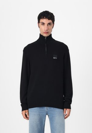 Pull en tricot noir avec un col montant et une fermeture éclair sur le devant. Présente une écusson logo carré sur la poitrine et des poignets et un ourlet côtelés.