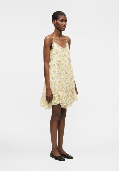 Γυναίκα που στέκεται με ένα αμάνικο, μέχρι το γόνατο, ανοιχτόχρωμο φόρεμα με floral σχέδιο και βολάν, φοράει μαύρες μπαλαρίνες σε απλό λευκό φόντο.