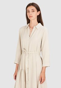 Beżowa sukienka zapinana na guziki z kołnierzykiem, długimi rękawami, ściągniętą talią z przewiązanym paskiem oraz marszczonym dolnym hemem. Gładki, lekki materiał.