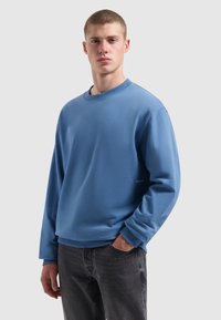 Jonge man met kort haar, gekleed in een blauwe longsleeve sweatshirt en zwarte jeans, staand met één hand in zijn zak tegen een effen achtergrond.
