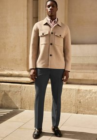 Jachetă din lână bej cu două buzunare frontale, butoane închise; combinată cu pantaloni gri închis și pantofi loafers negri. Textură fină, croi adaptat.