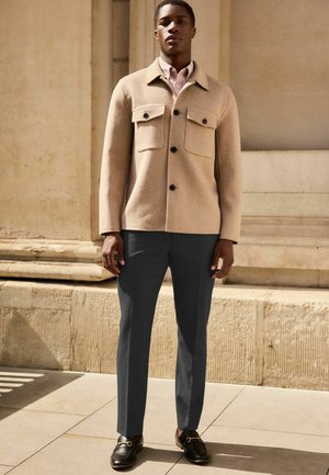 Chaqueta de lana beige con dos bolsillos frontales, botones oscuros; combinada con pantalones gris oscuro y mocasines negros. Textura suave, ajuste entallado.
