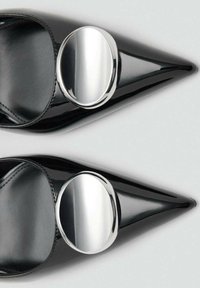 Mules de tacón alto de charol negro con punta puntiaguda, que presentan un gran adorno circular plateado en el lateral, textura lisa y acabado brillante.