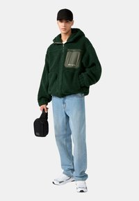 Zelená fleecová bunda s kapucňou, predným zipsom a náprsníkovou vreckom. V kombinácii so svetlo modrými džínami a bielymi teniskami. Čierna taška držaná v ruke.