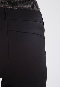 Pantalons extensibles noirs avec une texture lisse et une taille haute, présentant des détails de couture subtils et un accent métallisé à la taille.
