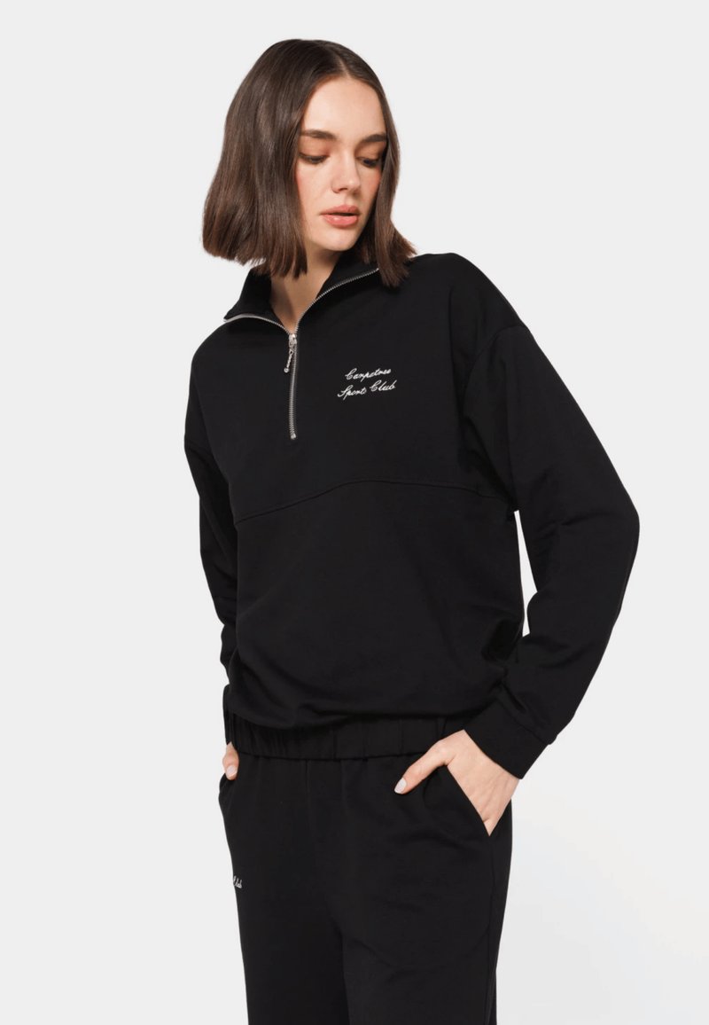 Kvinde iført en sort sweatshirt med halv lynlås og matchende sorte bukser med hænderne i lommerne, kigger ned mod sin venstre side.