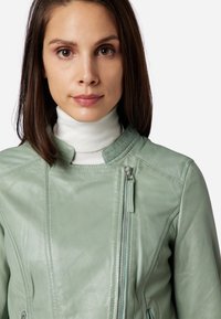 Mintgrön läderjacka med hög krage, sned dragkedjeslutning och slät yta. Innehåller en dragkedjeficka på bröstet.
