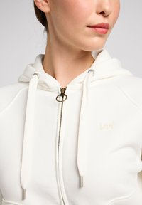 Weißer Kapuzenpullover mit Reißverschluss, verstellbaren Kordeln und gesticktem "Lee"-Logo. Aus weichem Stoff und mit silbernen Hardware-Details.