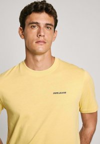 Geltona medvilnės marškinėliai su apvaliu kaklu ir trumpomis rankovėmis. Kairėje krūtinės pusėje yra mėlynas "PEPE JEANS" logotipas. Lygus paviršius.