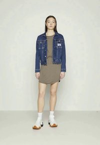 Jovem mulher em pé, vestindo um vestido casual às riscas, uma jaqueta de ganga azul, meias brancas e ténis brancos com solas de borracha.