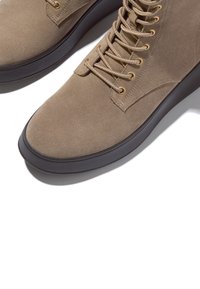 Beige suède enkellaarsjes met vetersluiting, gouden veterogen en een zwarte rubberen zool. Gladde textuur en afgeronde neus.