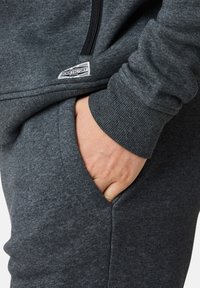 Grå sweatshirtmaterial med en jämn textur, med en sidficka och en etikett som läser "DENIM FACTORY." Dragkedjedetalj är synlig.