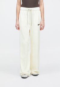 Személy laza, krémszínű Nike melegítőnadrágot visel, húzózsinórral a derékrészen, szürke Nike papuccsal, fehér háttér előtt.