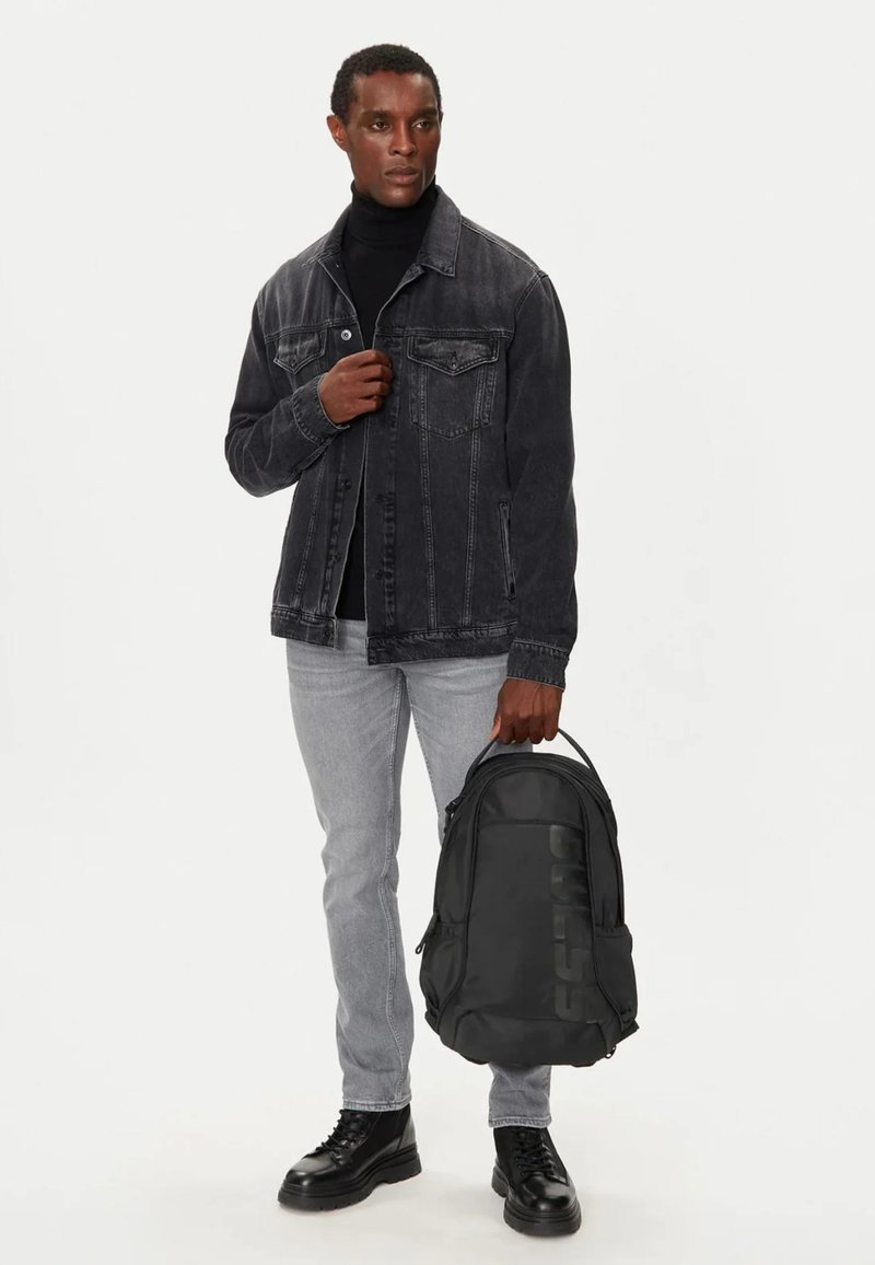 Giacca in denim nera con cuciture a vista, dolcevita nera, jeans grigi e zaino nero con logo in rilievo; robusti stivaletti neri con lacci.