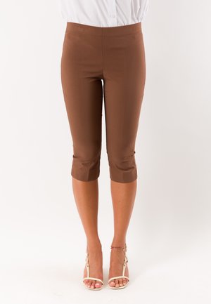 Donna con pantaloni capri marroni e sandali bianchi con tacco alto, in piedi davanti a uno sfondo bianco.