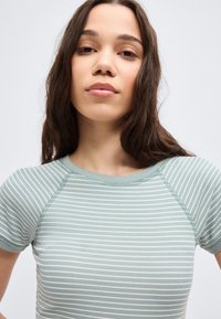 Νεαρή γυναίκα με μακριά καστανά μαλλιά, φορώντας κοντομάνικη ριγέ μπλούζα σε ανοιχτό πράσινο και λευκό, στραμμένη προς τα εμπρός μπροστά σε απλό φόντο.