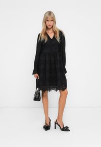 Eine Frau trägt ein schwarzes Spitzenkleid mit langen Ärmeln, schwarze Slingback-Pumps mit Schleifen und hält eine schwarze Handtasche. Sie steht vor einem weißen Hintergrund.
