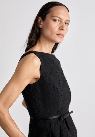 Svart ärmlös klänning med texturerad tyg, figursydd överdel och midjeband. Enfärgad, knälång, strukturerad design med subtila detaljer.