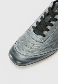Lædersneaker med en metallisk grå finish, der har en rund tå, sorte snørebånd, tekstureret syning og en glat, blank sektion ved tåen.