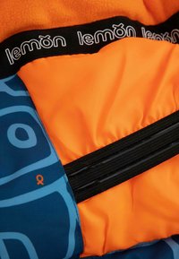 Kurtka z pomarańczowym polarem wewnątrz, niebieskim wzorzystym materiałem oraz czarną elastyczną taśmą z logo. Posiada dwa czarne zamki błyskawiczne.