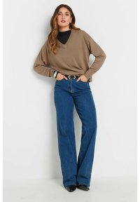 Brauner V-Ausschnitt-Pullover mit langen Ärmeln, kombiniert mit hoch taillierten blauen Schlagjeans. Silberner Gürtelakzent mit schwarzen spitzen Schuhen.