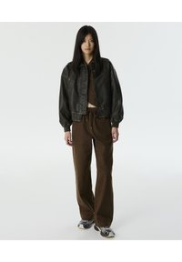 Chaqueta bomber de cuero negro con puños acanalados; pantalones de pana marrones; zapatillas con estampado. Silueta simple y moderna. Fondo neutro.