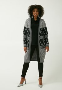 Larga chaqueta gris con acentos negros en patrón, que presenta diseños geométricos. Llevada sobre una blusa negra, combinada con pantalones negros y tacones plateados.
