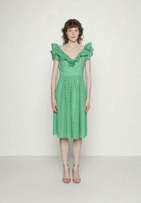 Vestido verde com bolinhas com mangas com babados e cintura justa, apresentando uma saia até aos joelhos. Usado com sandálias de tiras douradas.