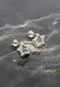 Sötliga stjärnformade örhängen i silver med klara ädelstenar, med en polerad finish och en studsback på en mörk, texturerad yta.