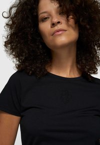 Mujer con cabello rizado que lleva una camiseta negra con un pequeño diseño en relieve sutil en la parte superior del pecho.