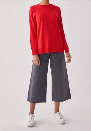 Person trägt einen knallroten Langarmpullover, dunkle weite cropped Jeans und weiße Sneakers, steht vor einem schlichten Hintergrund.